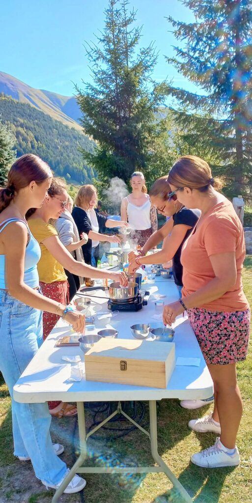 Mensen rond een tafel in de open lucht, bezig met de voorbereiding van een workshop voor het maken van voorwerpen of verzorgingsproducten, tijdens de Hygge-week – Welzijn in La Toussuire.