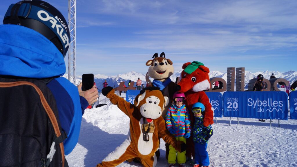 Photo prise de jour, pendant l'hiver en format paysage. Nous pouvons voir une personne de dos en train de prendre une photo de ses enfants qui posent avec les mascottes des stations des Sybelles lors du festi'kids. 