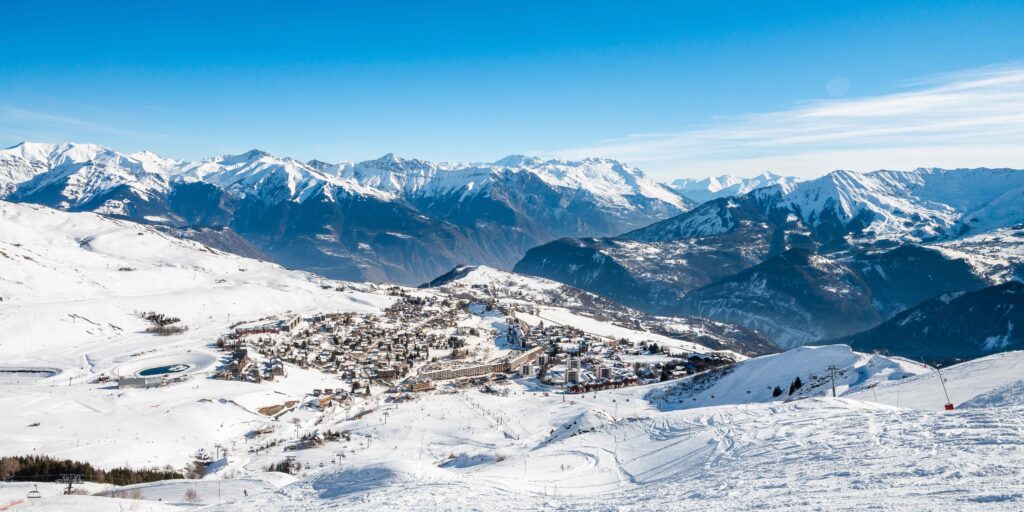 Foto overdag in liggend formaat. We zien het skioord La Toussuire in de winter. Het resort is bedekt met sneeuw. De foto is genomen vanaf de piste. Op de achtergrond zien we alle bergtoppen die het resort omringen. Ze zijn allemaal bedekt met sneeuw.
