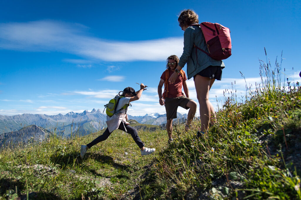 Photo en format paysage, prise de jour en été. Nous pouvons voir une famille de 3 personnes en pleine randonnée. Les deux parents sont tournés vers la fille qui est en train de sauter par dessus un petit fossé. En arrière plan nous voyons les montagnes avec les Aiguilles d'Arves.