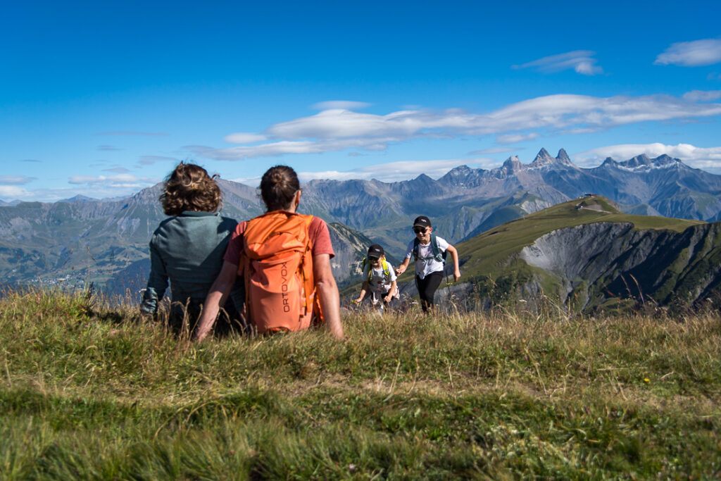 Photo prise en format paysage. La photo a été prise de jour en été. Nous pouvons au premier plan un couple assis dans l'herbe au sommet d'une montagne. Ils sont dos à l'objectif. Ils regardent leurs deux filles qui courent vers eux. En arrière plan nous pouvons voir les Aiguilles d'Arves.