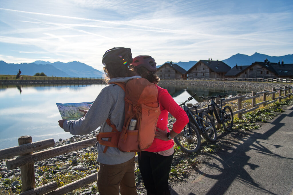 Photo prise de jour en été, en format paysage. Nous pouvons voir un couple de vététiste au premier plan en train de regarder une carte pour choisir leur itinéraire. Leurs vélos sont posés juste à côté d'eux sur une barrière. Ils se trouvent à proximité d'un lac. Au second plan nous voyons le lac avec en arrière plan des chalets.