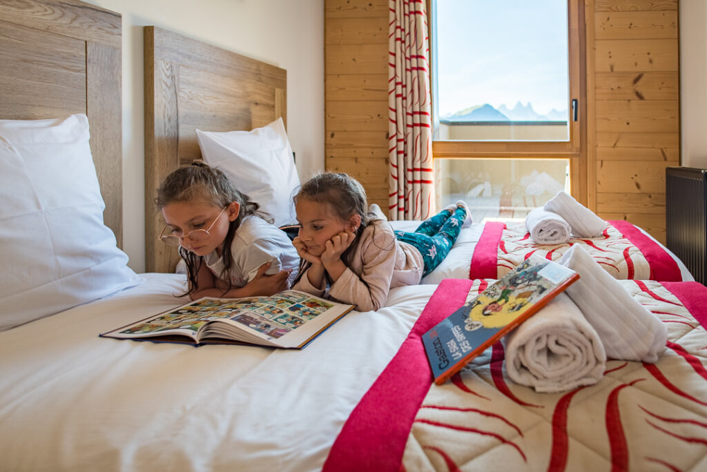Photo en format paysage prise de jour. Nous pouvons deux filles en train de lire une BD sur un lit.