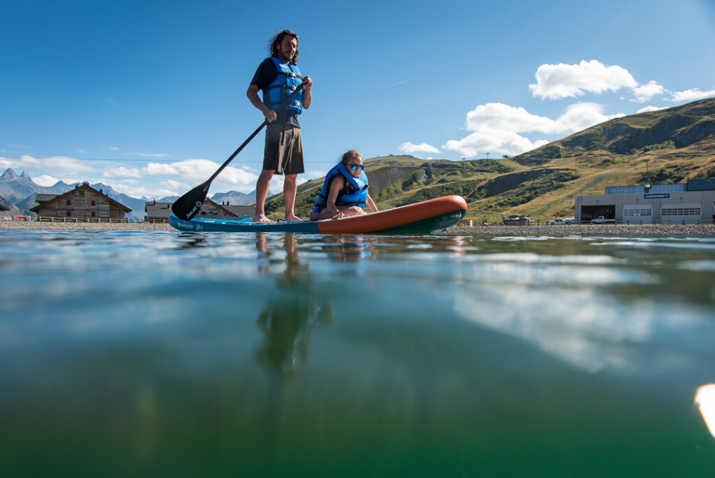 Photo prise de jour en été en format paysage. Nous pouvons observer un père et sa fille en train de faire du paddle, sur le lac de La Toussuire. La fille est en position assise tandis que le père est debout derrière elle. En arrière plan nous voyons les montagnes de La Toussuire