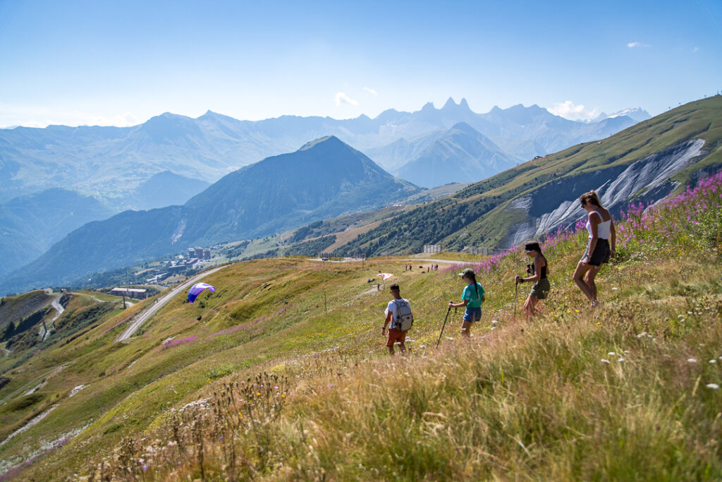 Hikers in summer at La Toussuire, walking along a flower-filled path with views of the mountains and the Aiguilles d'Arves.