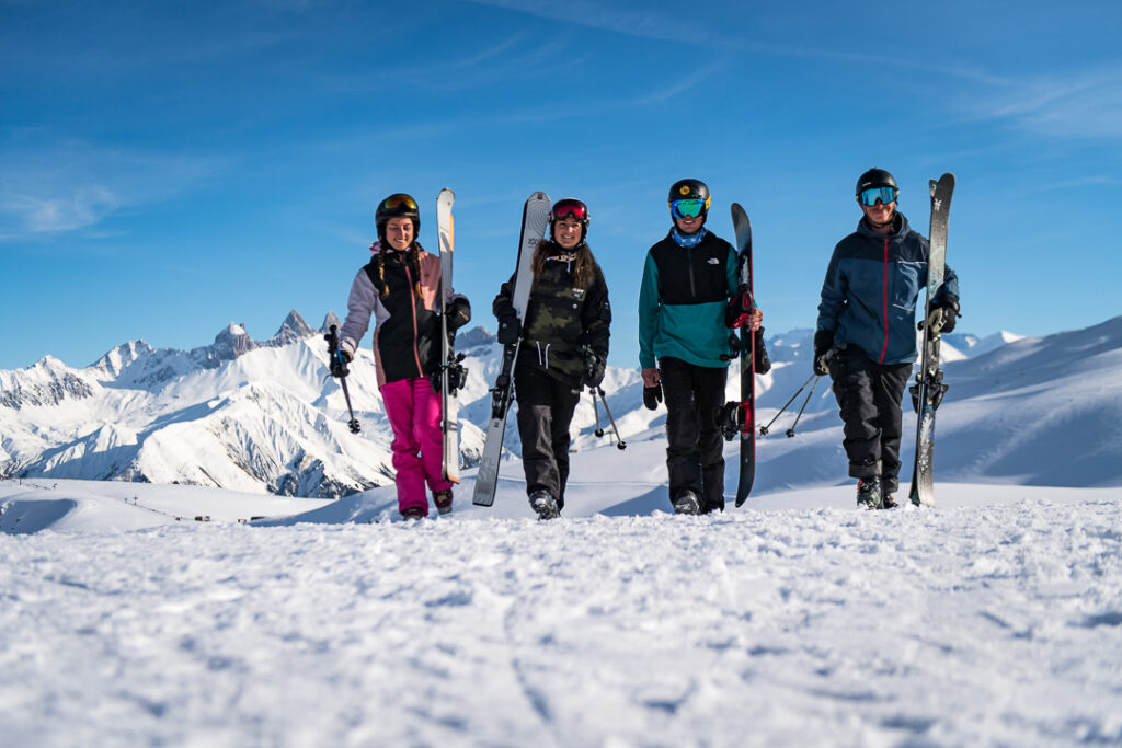 Photo prise de jour en format paysage en hiver. Nous pouvons voir un groupe d'amis en train marcher dans la neige les uns à côté des autres en portant leurs matériels de ski. 