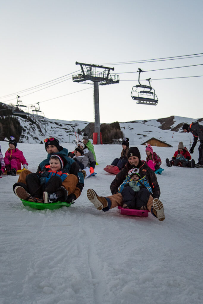 Photo prise au crépuscule en format portrait. La photo a été prise pendant l'hiver. Nous pouvons voir une famille en train de faire une descente de luge. Chaque parent est avec un enfant sur la luge. 