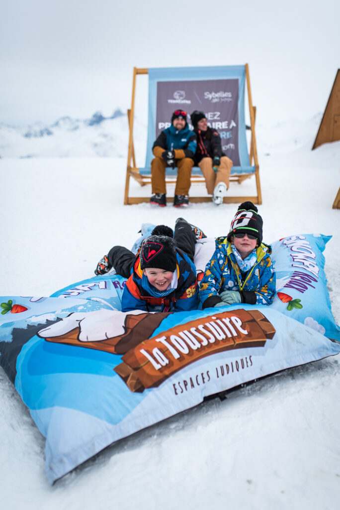 Photo prise en format portrait de jour. La photo a été prise en hiver. Nous pouvons voir une famille en train de passer du bon temps à l'espace de loisir de Snowpy Mountain. Au premier plan, deux enfants sont allongés sur un coussin géant qui est posé sur la neige. Au second plan nous pouvons voir les parents assis dans un transat géant. 