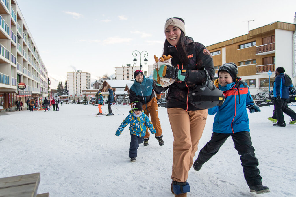 Photo prise de jour en format paysage pendant l'hiver. Nous pouvons voir une famille courir dans la neige. La mère est devant en tenant un sachet de churros, les enfants lui courent après pour récupérer les chichis.