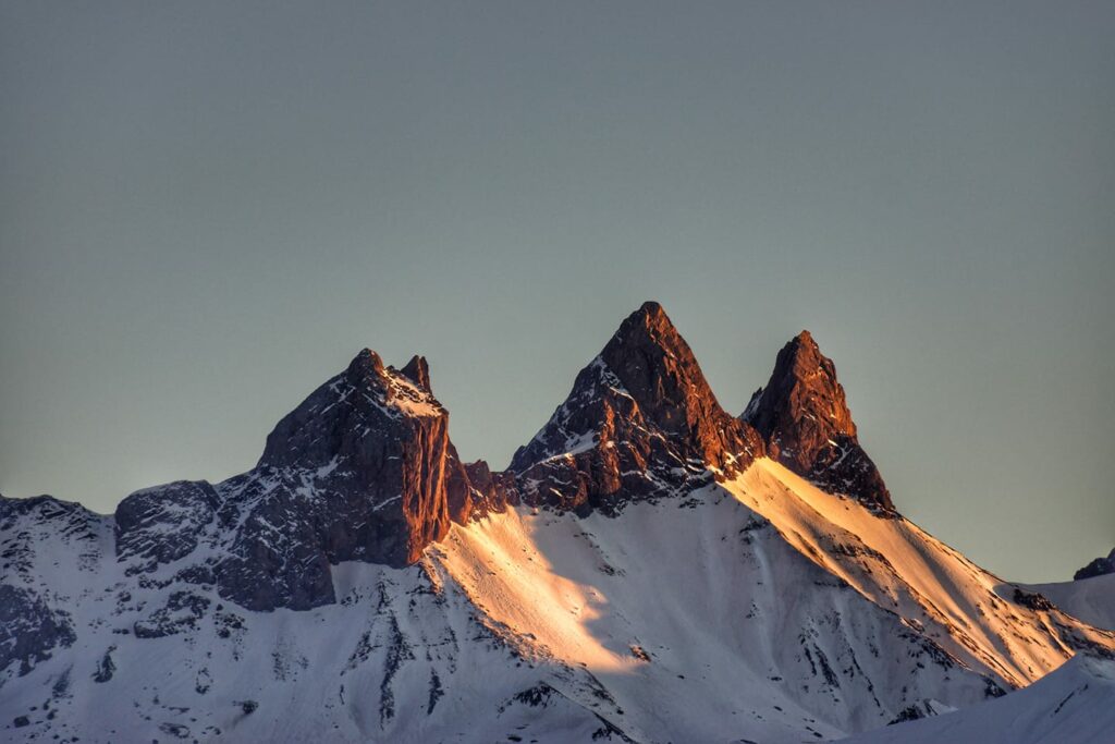 Landschapsfoto genomen bij zonsondergang. We zien de besneeuwde Aiguilles d'Arves bij zonsondergang met de oranje stralen die weerkaatsen op de rotsen en de sneeuw.