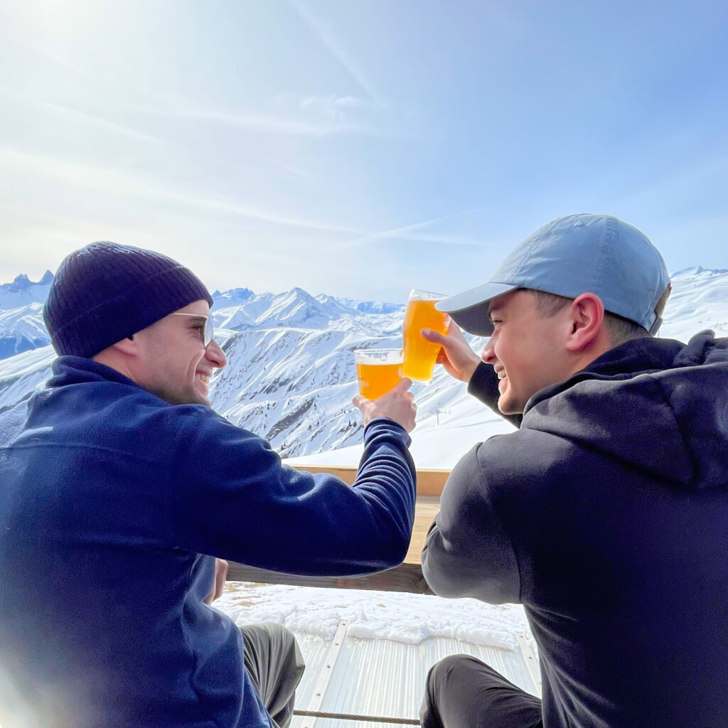 Photo prise de jour en format portrait en hiver. Nous pouvons voir de amis prenant un verre en terrasse sous le soleil de La Toussuire. Ils sont sur la terrasse d'un restaurant d'altitude, avec au second plan les sommets enneigés.