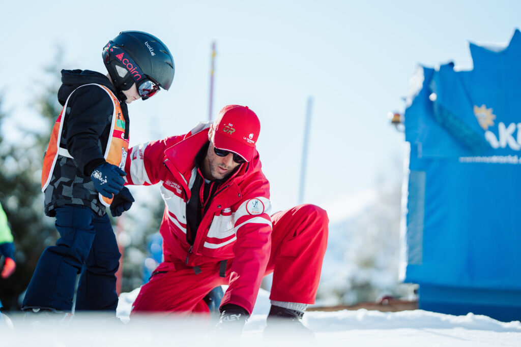 Landscape photo taken during the day in winter. We can see a ski instructor giving a ski lesson to a child. 