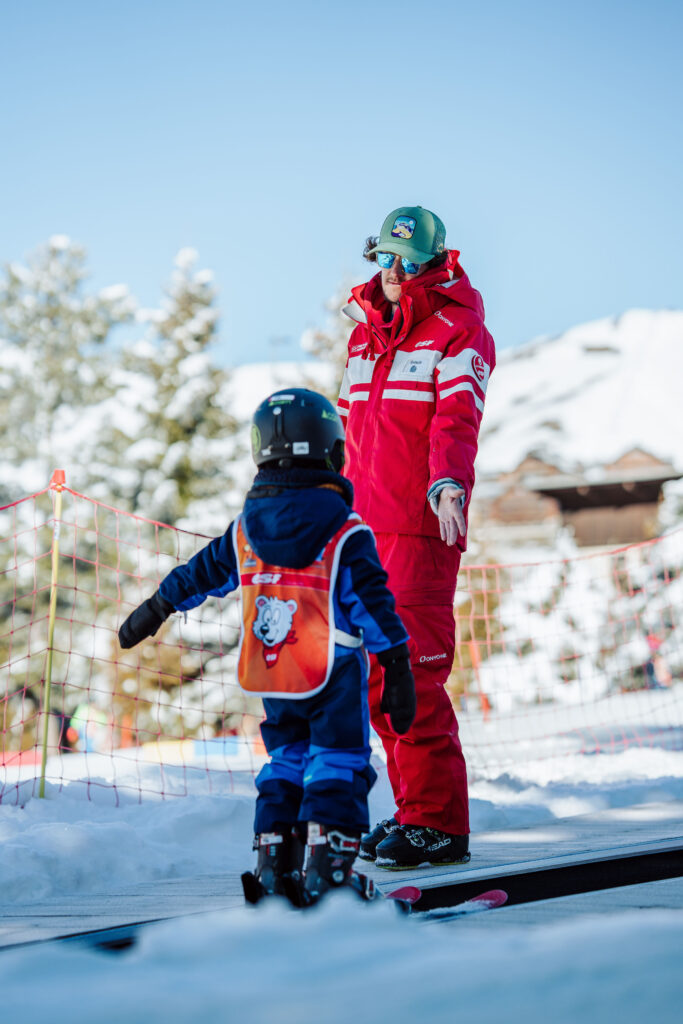 Portrait shot taken during the day in winter. We can see a ski instructor giving a lesson to a child. 