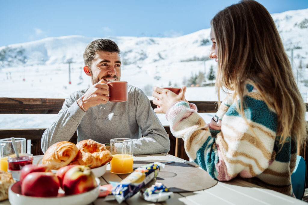 Overdag genomen in liggend formaat, zien we een koppel ontbijten op het terras van hun flat. Ze hebben elk een kopje in hun hand. Op de achtergrond zien we de besneeuwde bergen.