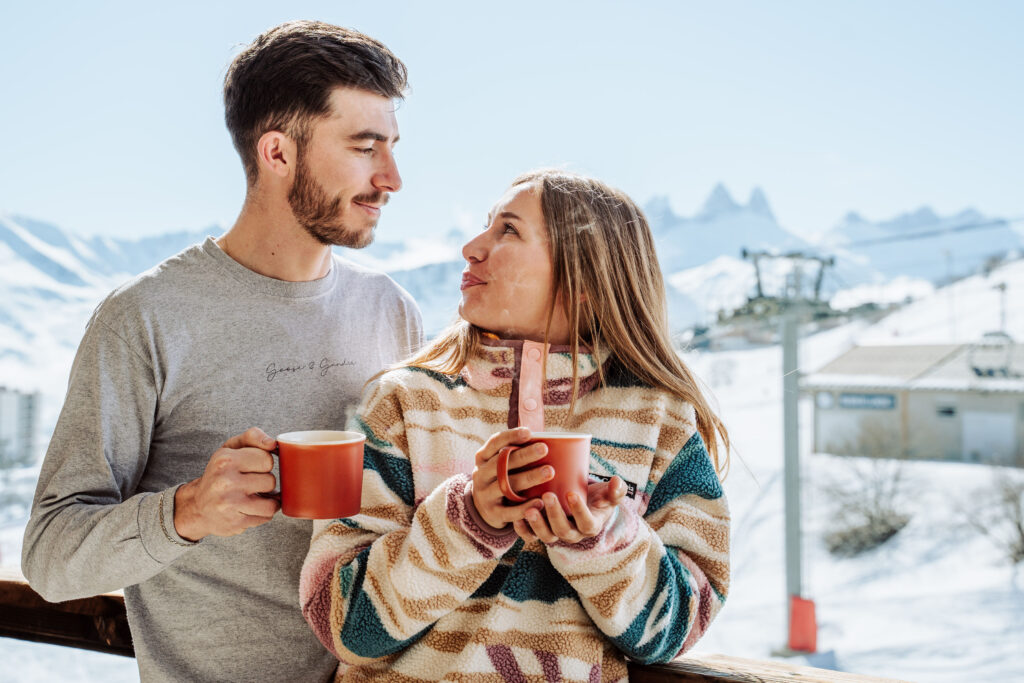 Daytime landscape shot in winter. We can see a couple with a cup of coffee in their hand looking into each other's eyes. In the background we can see a ski lift and snow-capped mountains.  