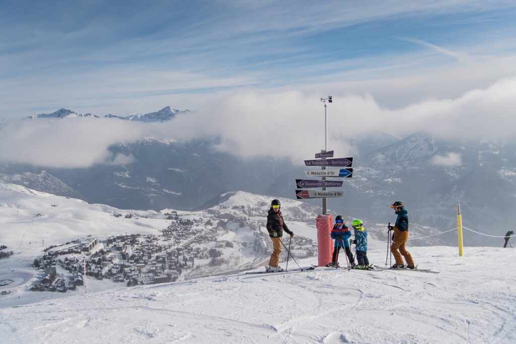 Foto overdag in horizontaal formaat. De foto is genomen in de winter in het skigebied waar we een familie skiërs hun volgende afdaling zien kiezen. Op de achtergrond zien we het resort La Toussuire en de besneeuwde bergen.