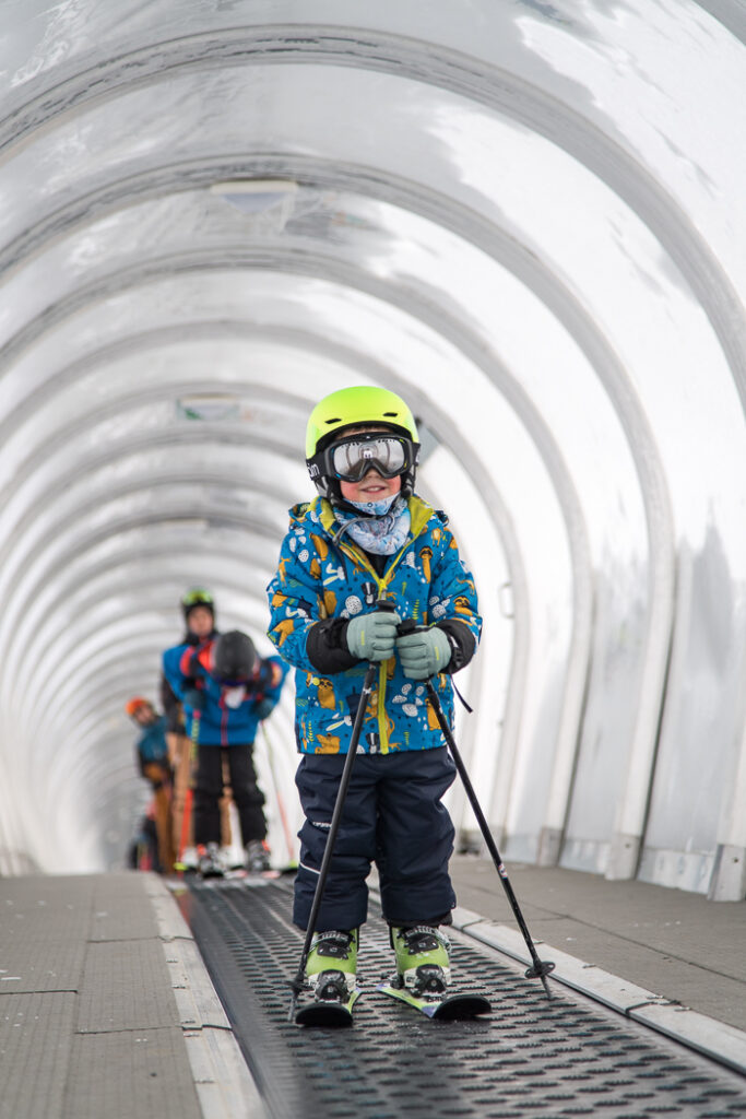 Photo au format portrait avec un enfant en tenue de ski sur un tapis tube pour débutant. L'enfant est debout équipé de la tête aux pieds avec un grand sourire