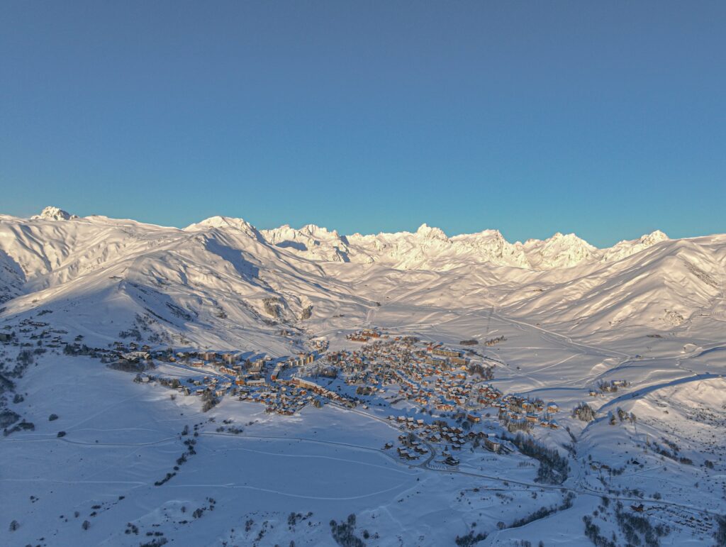 Photo prise lors d'un levé de soleil. Il s'agit d'une vue en drone en contre plongeur la station en pleine hiver avec un grand ciel bleu et le domaine skiable 