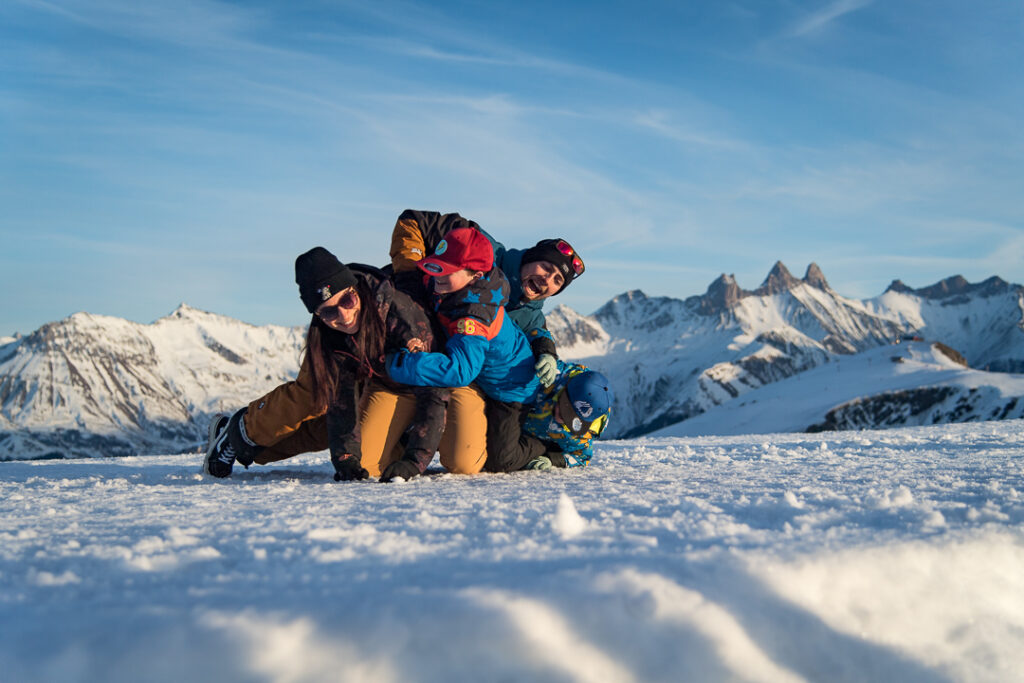 Photo prise en hiver lors d'une fin de journée. La photo est en horizontal et nous pouvons voir au premier plan une famille en train de jouer avec la neige et au second plann un couché de soleil sur les aiguilles d'arves