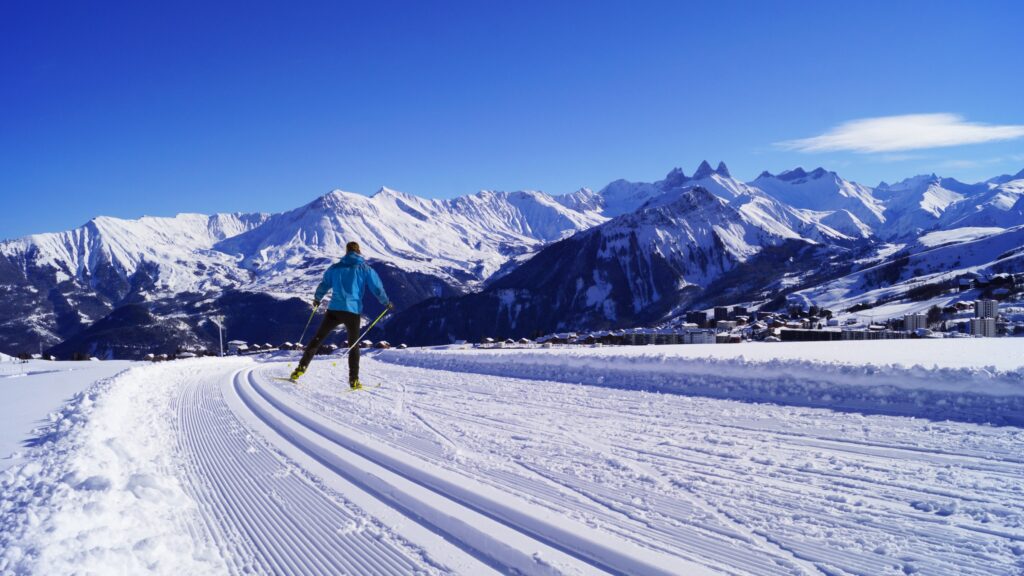 Photo prise sur les pistes de ski de fond de la Toussuire. Nous pouvons voir un skieur en action de dos avec les traces de skieurs au premier plan et un panorama montagneux en fond avec notamment les Aiguilles d'Arves.