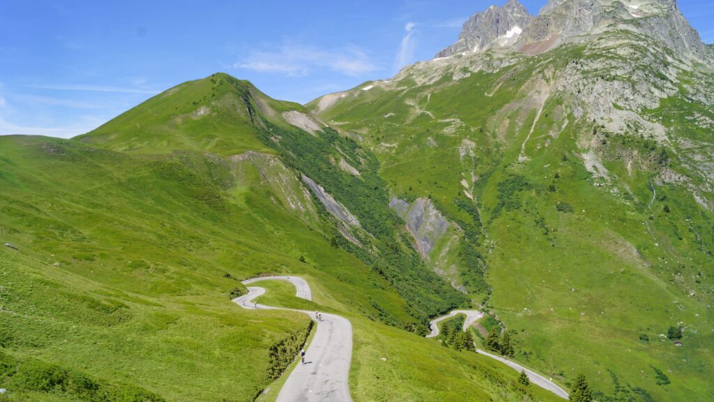 Vue en plongée sur une route de montagne sinueuse, entourée de vastes prairies vertes. Plusieurs cyclistes montent la route en lacets, qui serpente entre les pentes herbeuses. À l’arrière-plan, des sommets rocheux et escarpés se dressent sous un ciel bleu clair.