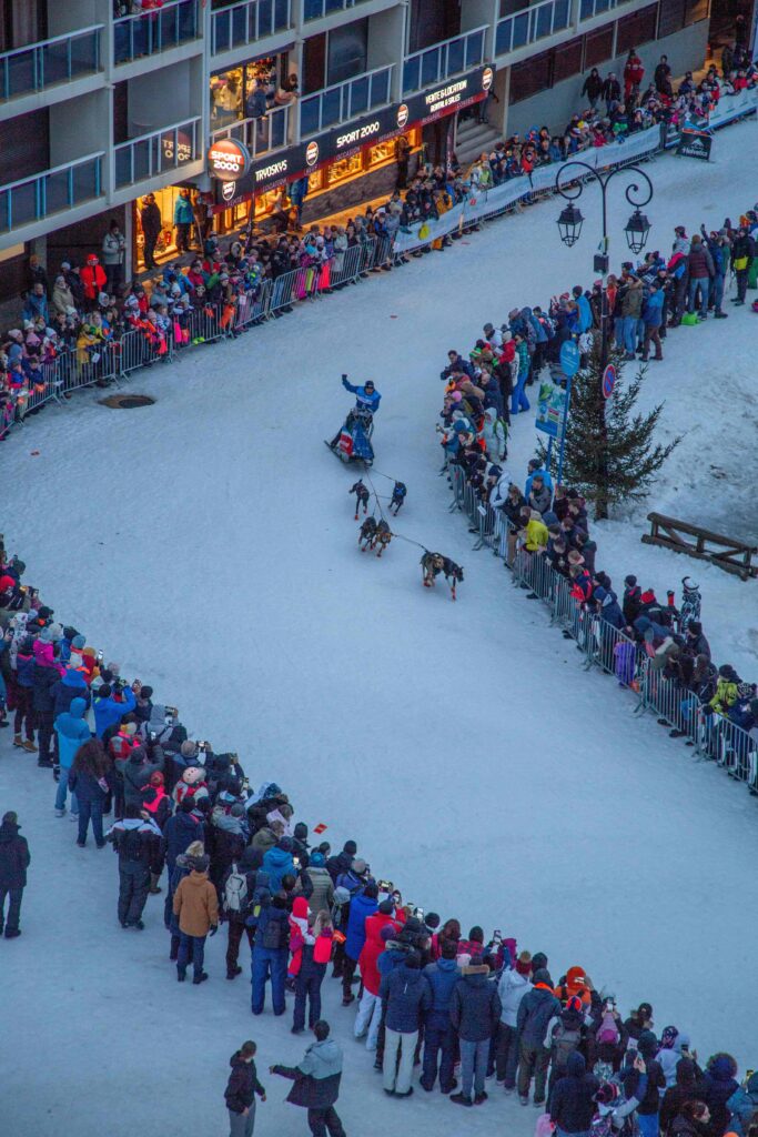 Photo prise en format portrait à la tombée de la nuit pendant l'hiver. La photo a été prise pendant l'événement la Grande Odyssée VVF qui est une course de chiens de traîneau. Nous pouvons voir un musher avec ton attelage. Il vient se lancer depuis la ligne de départ. La photo a été prise en hauteur de chaque côté du musher il y a des barrières avec une foule pour l'encourager. La