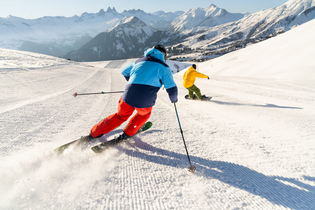 Photo prise de jour en format paysage en hiver. Nous pouvons voir un skieur et un snowboarder en train de descendre une piste en direction de la station de La Toussuire. Les deux personnes sont dos à l'objectif. 