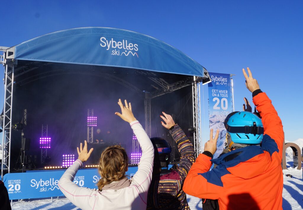 Photo prise de jour en format paysage, pendant l'hiver. Nous pouvons voir un groupe d'amis en train de danser pendant le concert d'ouverture organisé par le domaine skiable. Au second plan, nous pouvons voir la scène avec les artistes.