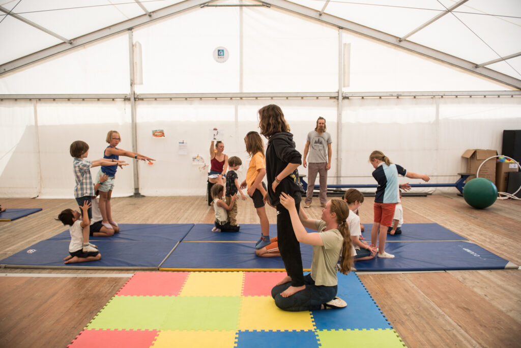 Photo en format paysage prise de jour. Nous pouvons voir des enfants en train de pratiquer un atelier cirque sur des portées. Les enfants sont par groupes de deux.