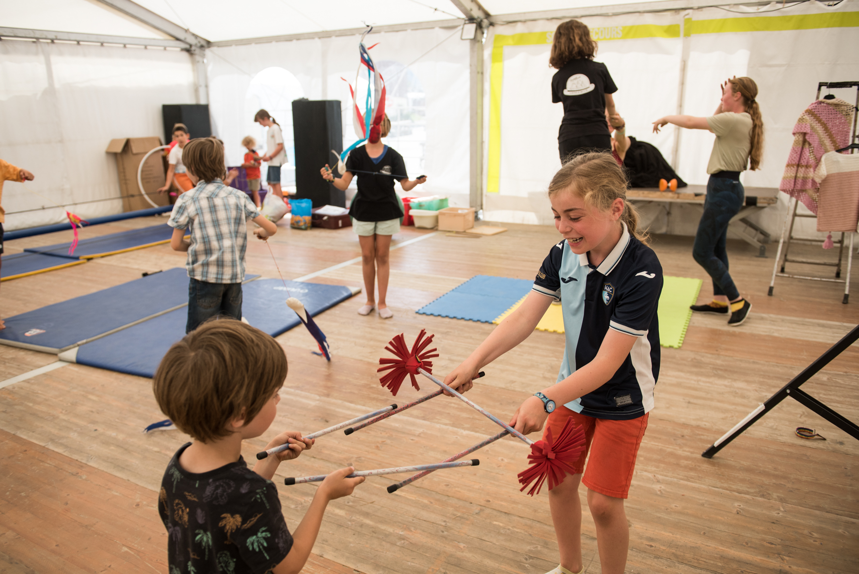 Photo prise en format paysage de jour. Nous pouvons voir au premier plan deux enfants en train de pratiquer un atelier jonglage. Autour d'eux nous pouvons voir d'autres enfants s'essayer à différents arts du cirque.