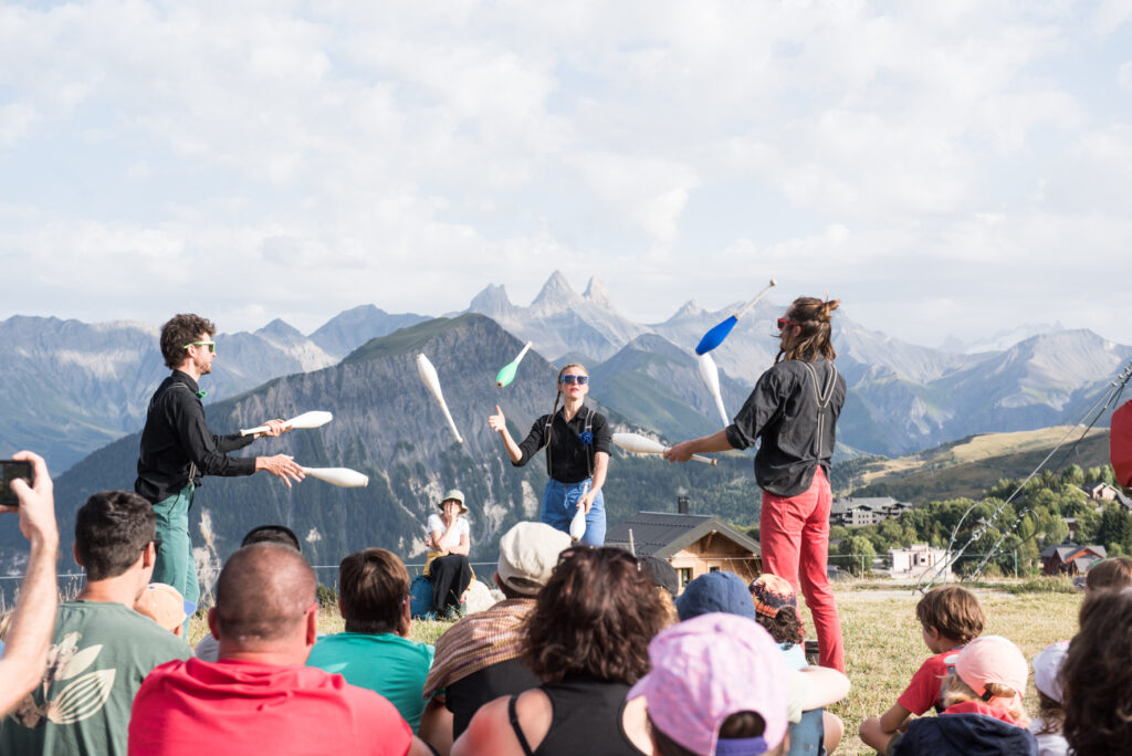 Photo prise de jour en format paysage. Nous pouvons voir trois personnes, deux hommes et une femme, en train de faire du jonglage devant un public avec en arrière plan le Mont Charvin et les Aiguilles d'Arves.