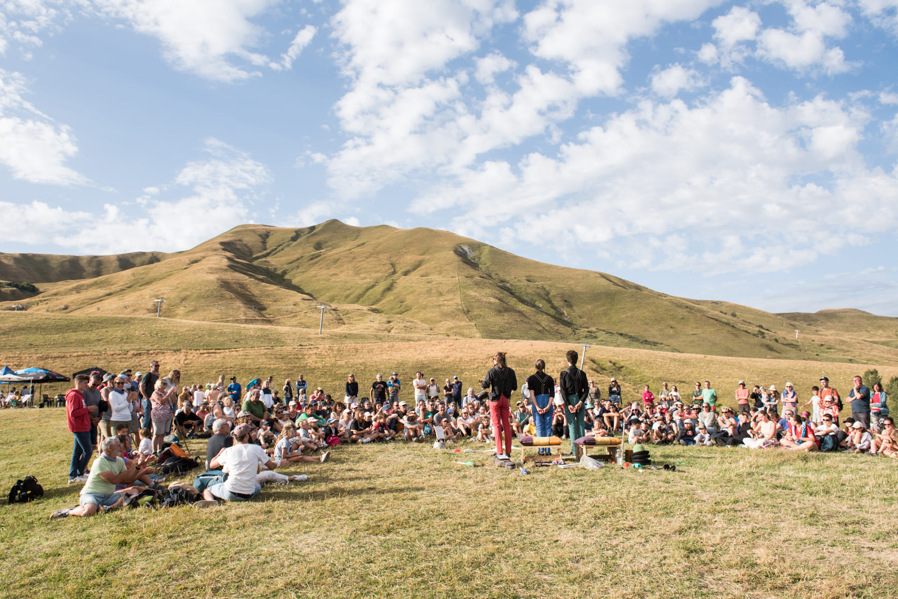 Photo prise de jour en été, en format paysage. Nous pouvons voir des personnes en train d'assister à un spectacle. Ils sont pour la plupart assis en arc de cercle dans l'herbe, certains d'autres eux sont débout. Ils regardent trois artistes qui sont debout face à eux. La photo a été prise dans le dos des artistes. En arrière plan nous pouvons observer la montagne du Grand Truc.