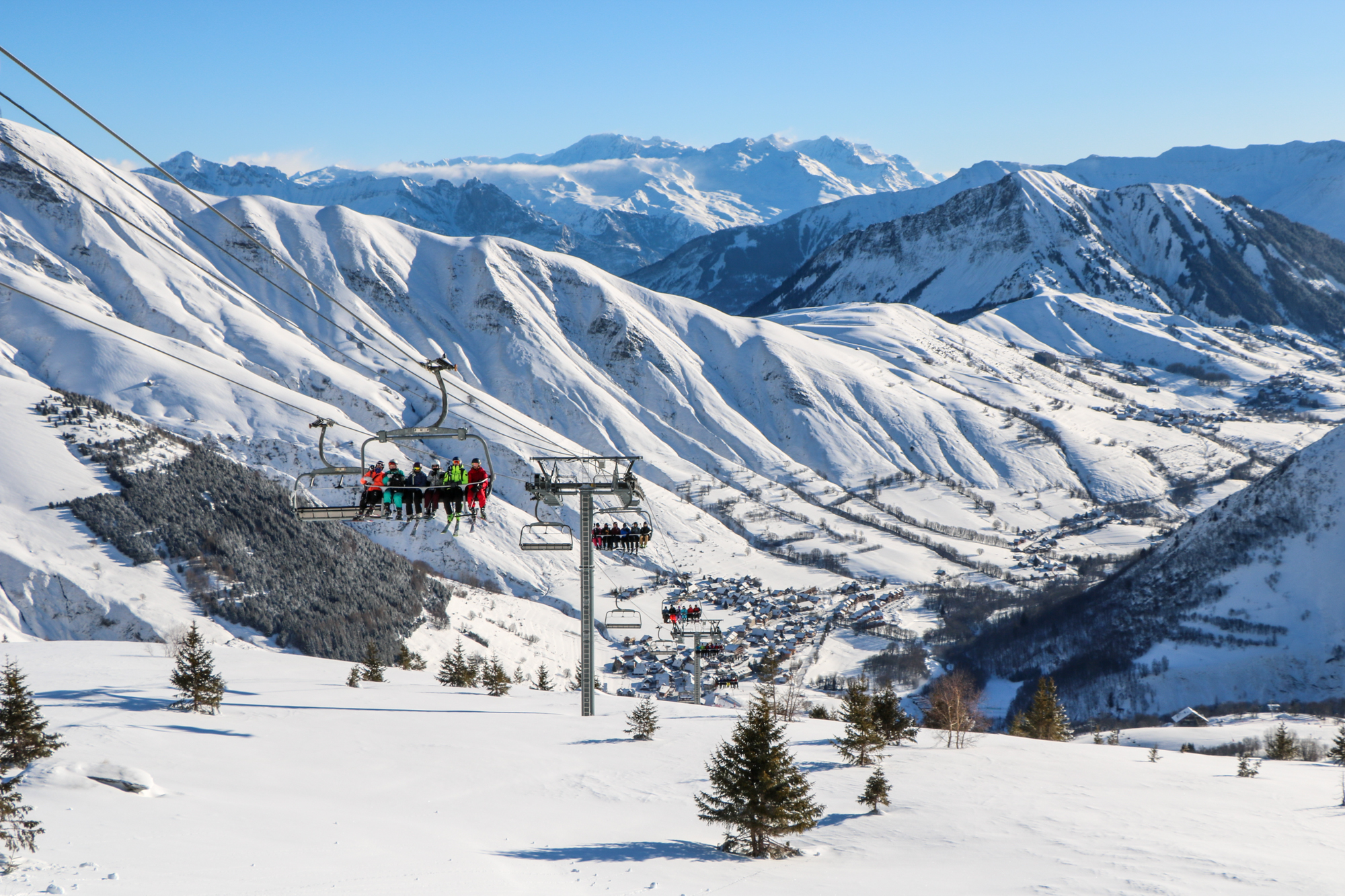 Photo en format paysage prise de jour. Nous pouvons voir la station de Saint Sorlin d'Arves en vue aérienne. La photo a été prise en hiver. Nous pouvons voir au premier plan un télésiège avec des skieurs dessus. Derrière la station de Saint Sorlin d'Arves en contre bas. En fond nous pouvons voir les montagnes enneigées.
