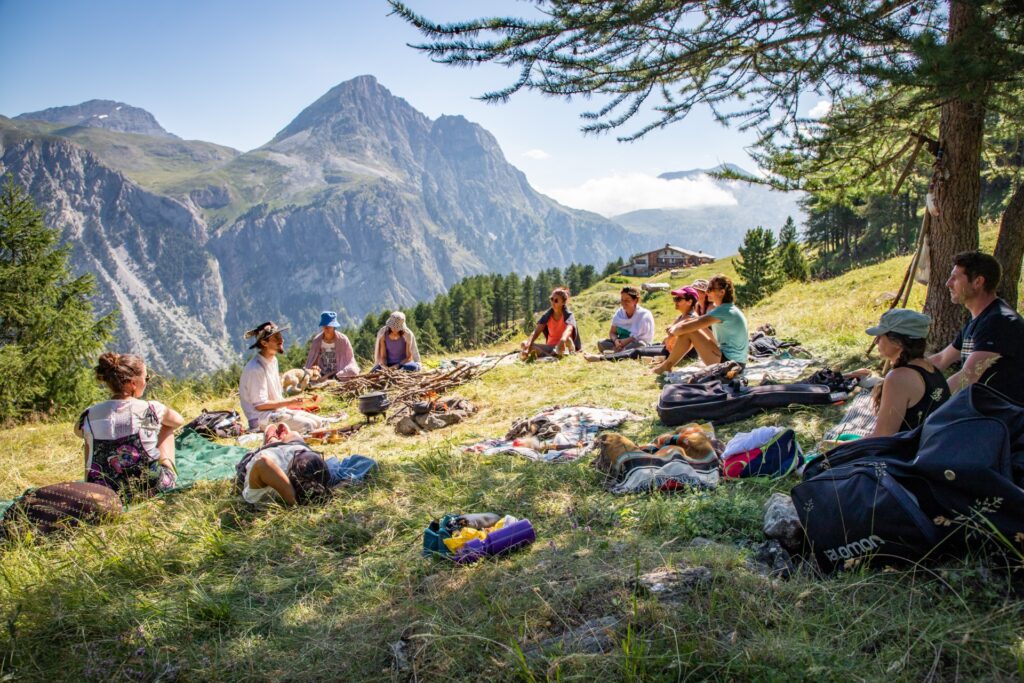 Groupe de personnes assis en cercle sur l’herbe, au milieu de la montagne, entourant un feu de camp improvisé, lors de la Semaine Hygge – Bien‑être à La Toussuire.