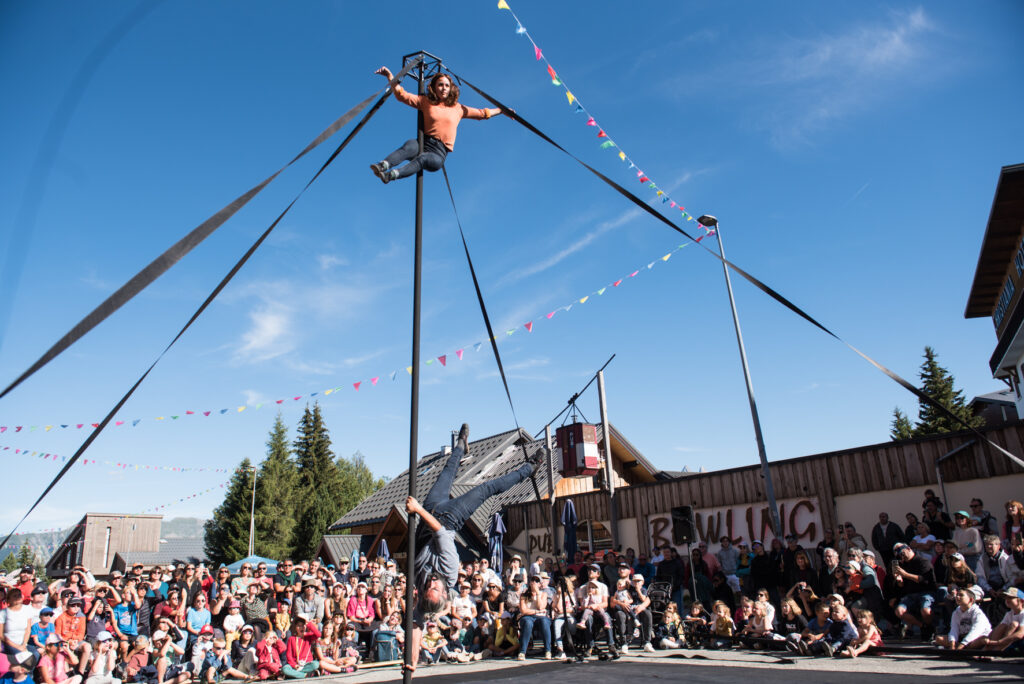 Photo prise de jour en format paysage en été. Nous pouvons voir un des spectacle du festival le Spectacle Est Dans La Rue. Nous pouvons voir deux artistes en train d'effectuer des figures dans les airs sur un poteau.