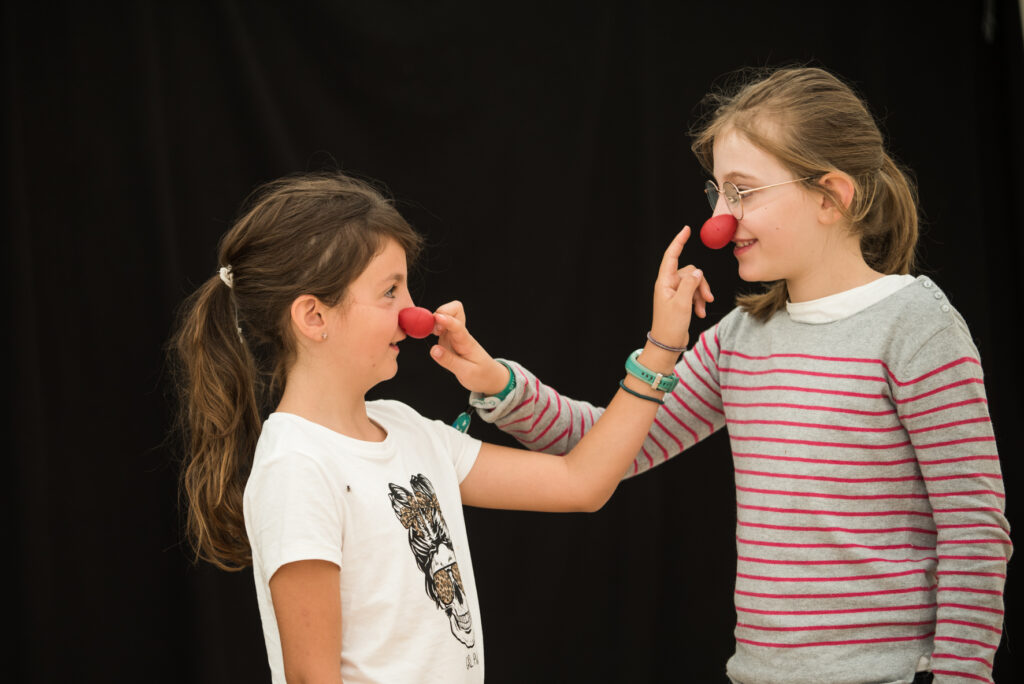 Photo prise de jour en format paysage. Nous pouvons voir deux jeune filles qui se font face à face. Elles portent toutes les deux un nez rouge qu'elles touchent mutuellement avec leur index.