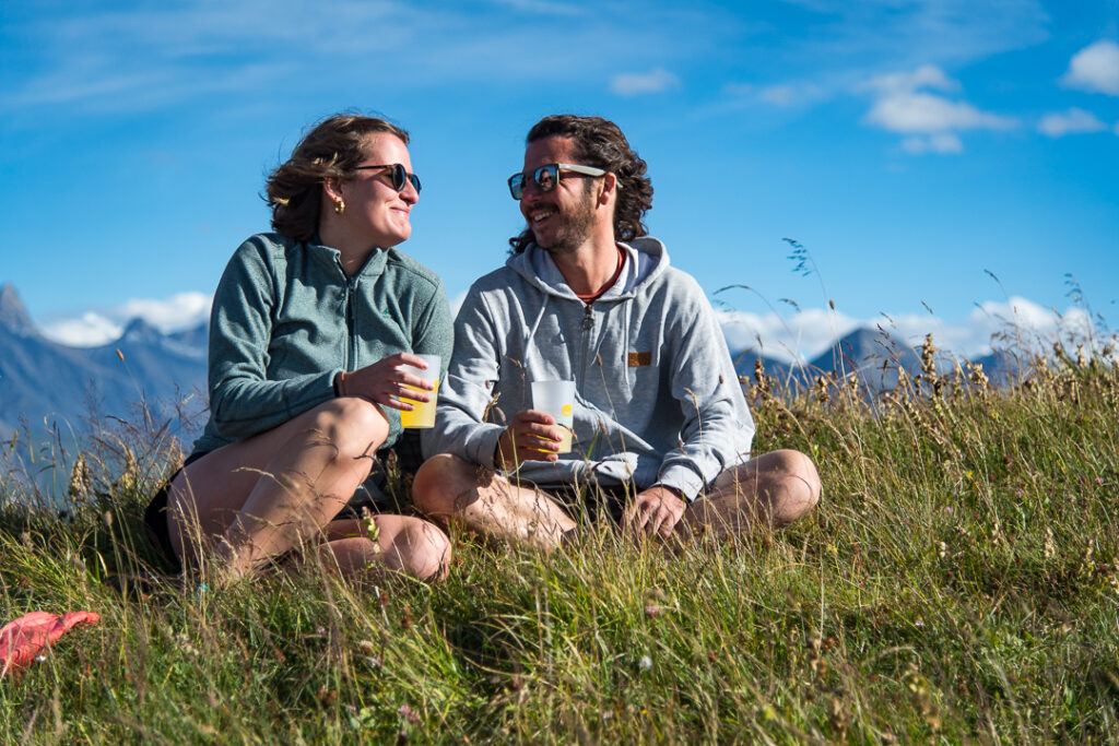 Photo prise de jour en format paysage. La photo a été prise en été. Nous pouvons voir un couple profiter d'une pause pour se désaltérer. Ils sont assis dans l'herbe avec chacun un verre dans la main, tout en se regardant