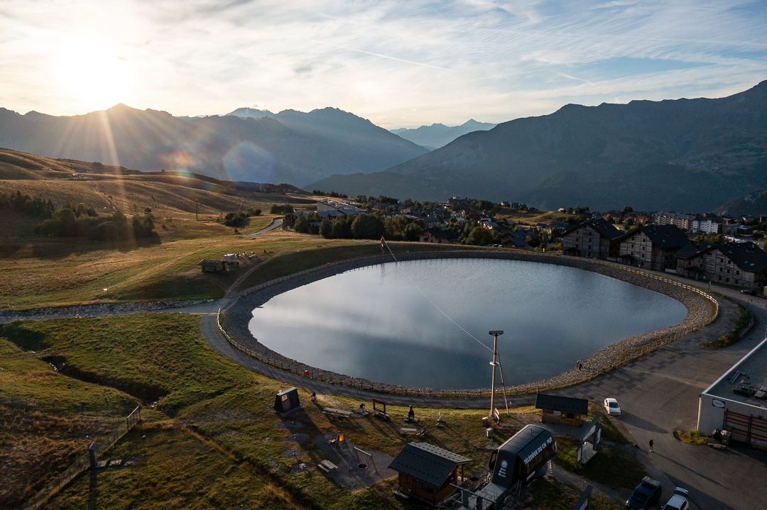 Photo en vu drone au lever du soleil. Nous pouvons voir le lac Eriscal avec la tyrolienne et l'espace ludique du Chaput. Au second plan des chalets avec les montagnes.