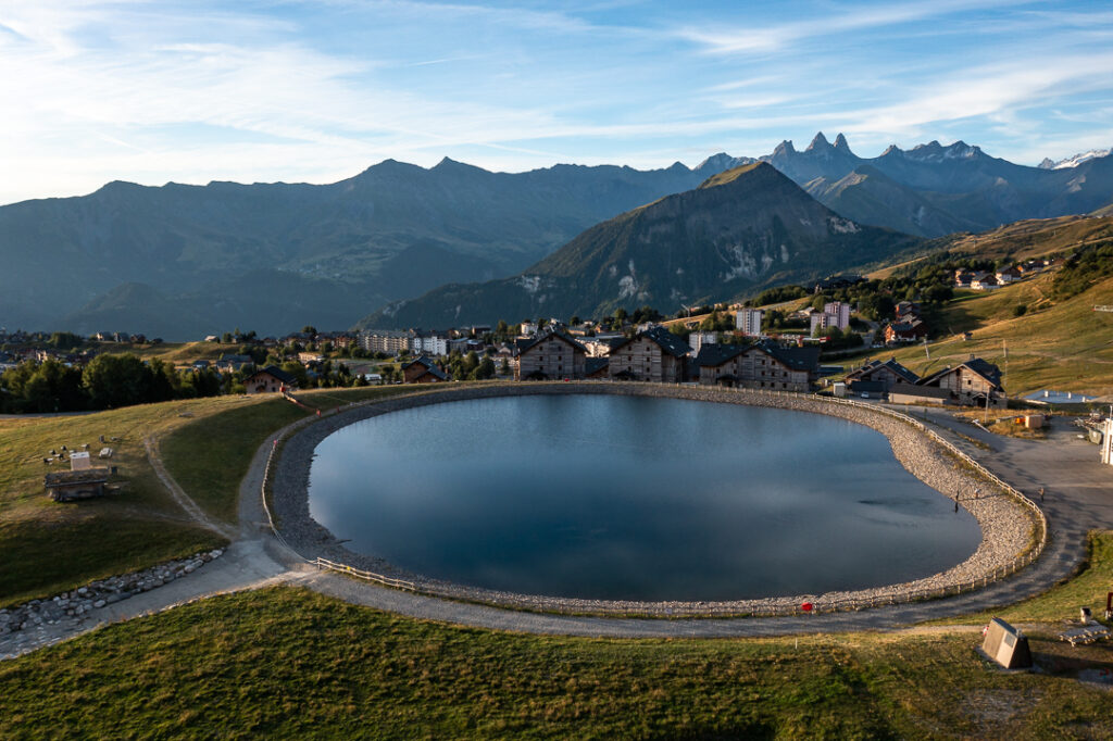 Photo en vu drone au lever du soleil. Nous pouvons voir le lac Eriscal. Au second plan une partie de la station de La Toussuire avec en arrière plan le Mont Charvin et les Aiguilles d'Arves.