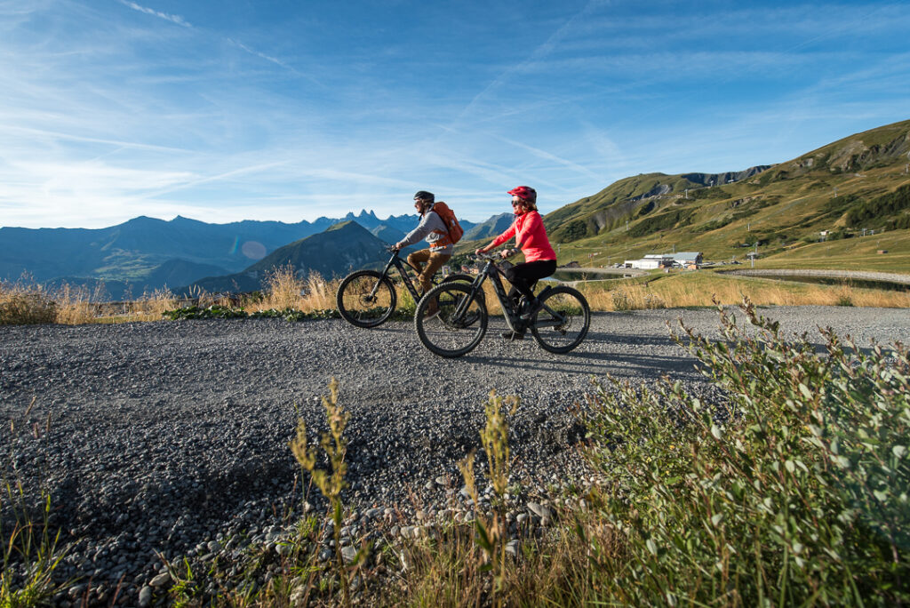 Photo prise de jour en été en format paysage. Nous pouvons voir un couple faire une randonnée VTTAE sur un chemin de terre sur La Toussuire. En arrière plan nous apercevons le Mont Charvin et les Aiguilles d'Arves