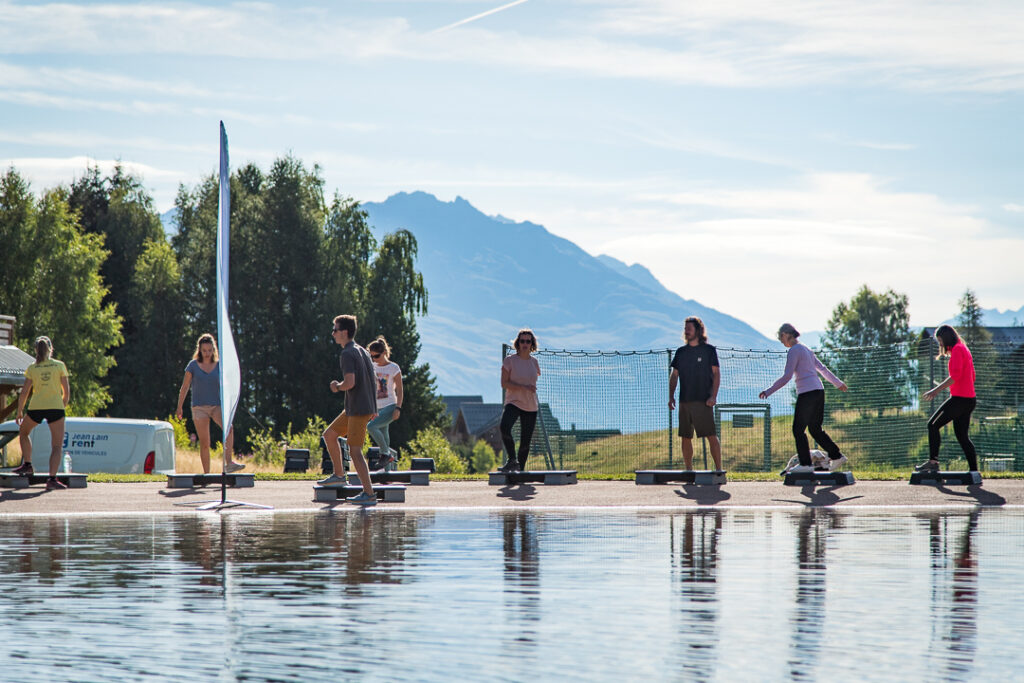 Photo prise de jour en été en format paysage. Nous pouvons voir des vacanciers faire l'animation "activité step" à proximité du plan d'eau de La Toussuire