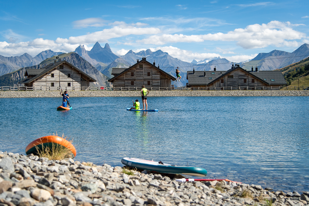 Photo prise de jour en été en format paysage. Au premier plan, nous pouvons deux paddle poser sur le bord du lac. Au second plan nous pouvons observer une famille en train de faire du paddle, sur le lac de La Toussuire. Au dessus d'eux une personne fait de la tyrolienne. En arrière plan nous voyons des chalets avec les Aiguilles d'Arves.