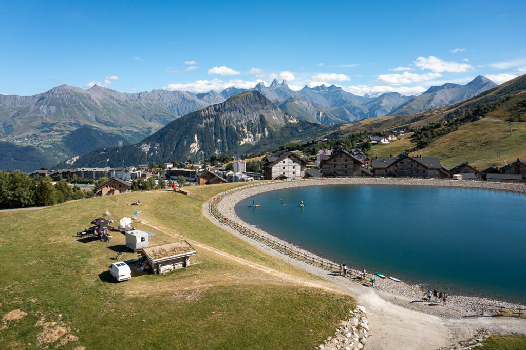 Photo prise en drone, de jour en été en format paysage. Au premier plan, nous pouvons voir le snack Chez bib avec la terrasse au bord du lac. Au second plan nous pouvons observer une famille en train de faire du paddle, sur le lac de La Toussuire. Au dessus d'eux une personne fait de la tyrolienne. En arrière plan nous voyons des chalets avec les Aiguilles d'Arves.