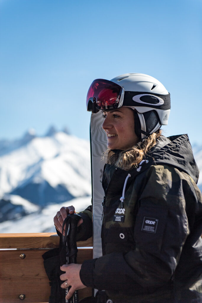 Photo prise en format portrait de jour en hiver. Nous pouvons voir une femme en tenue de ski portant un casque et un masque sur la tête et tenant ses skis. En arrière plan nous pouvons voir des montagnes enneigées.