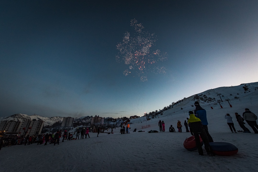 Photo prise en paysage au tombé de la nuit pendant l'hiver. un feu d'artifice est tiré. Des familles au pied des pistes regardent le spectacle.