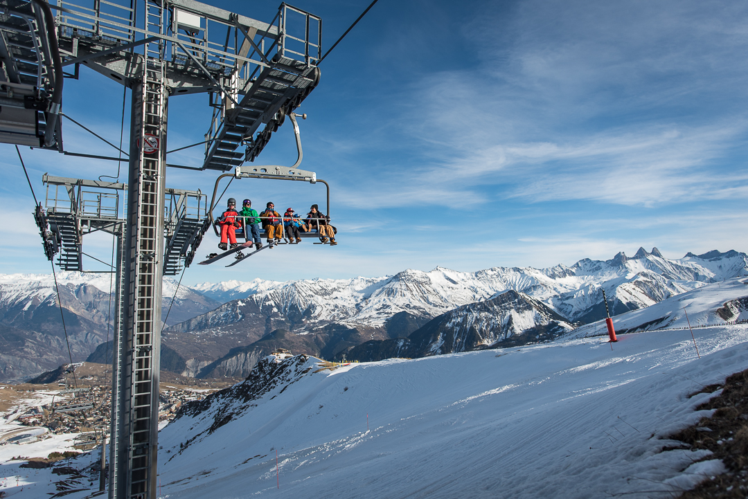 Photo en format paysage prise de jour. La photo a été prise en hiver. Nous pouvons voir 5 personnes sur un télésiège, prêt à aller skier.