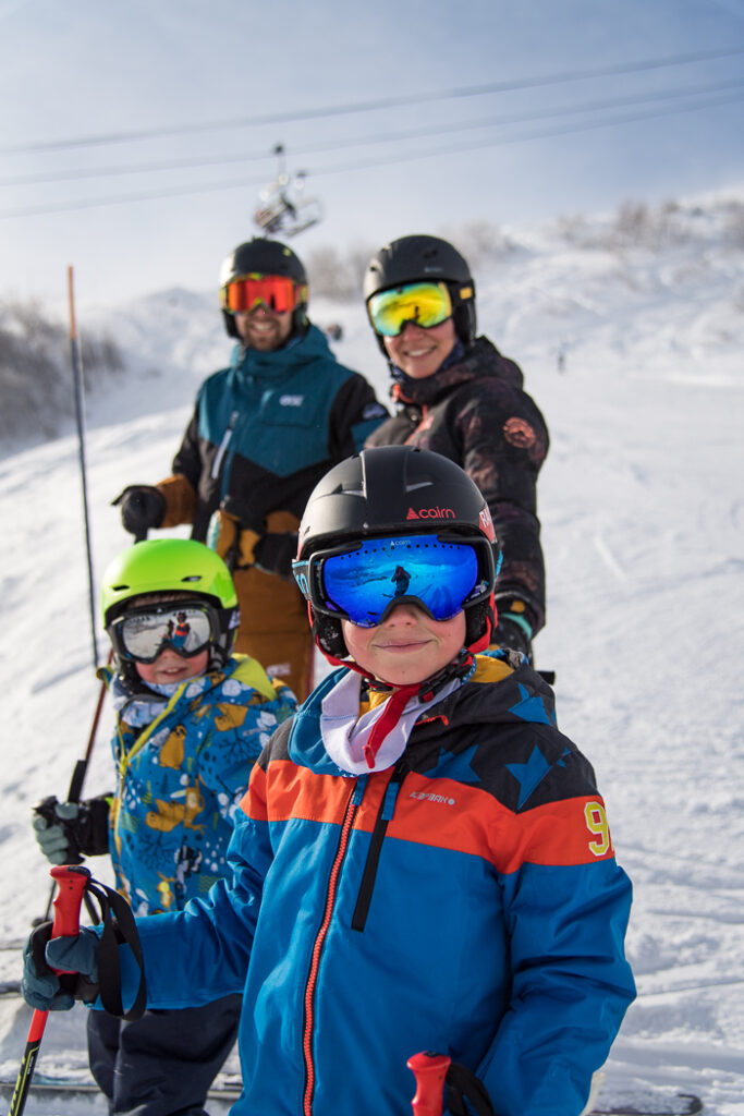 Photo prise en format portrait. Nous pouvons voir une famille composée de deux parents et deux enfants en train de se faire prendre en photo tous ensemble. Les enfants sont en premier plan les parents sont au second plan. Ils sont en train de faire une sortie ski.