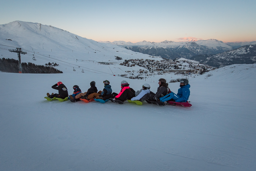 Photo prise en paysage, au crépuscule. Nous pouvons voir un groupe de personne en train de faire l'activité snakegliss. Ils sont 8 personnes au total. Chaque personne est assise sur une luge qui sont toutes reliées entre elles.
