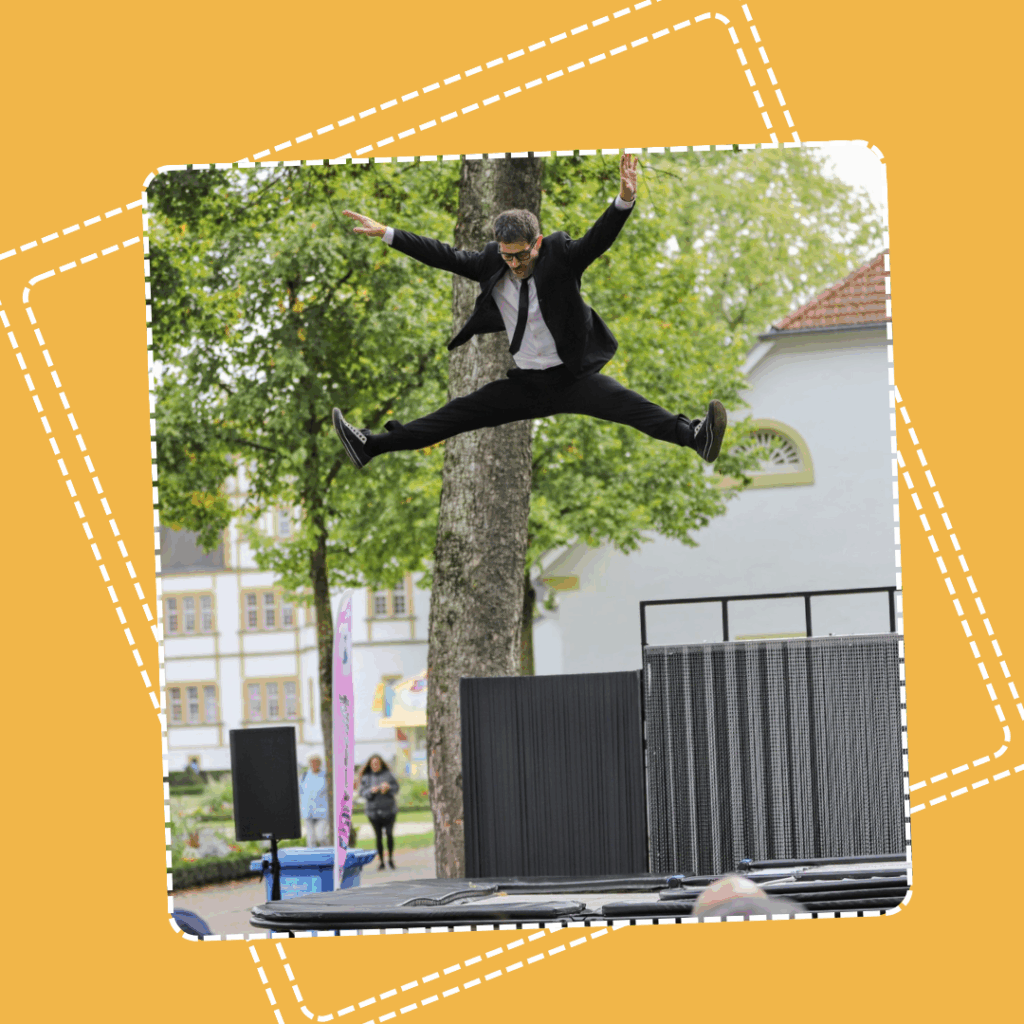Un artiste en costume noir et cravate saute en grand écart au-dessus d’un trampoline en plein air. Il est suspendu en l’air, bras tendus, dans un geste spectaculaire de voltige acrobatique. À l’arrière-plan, des bâtiments et des arbres évoquent un cadre urbain arboré, avec quelques spectateurs en retrait.