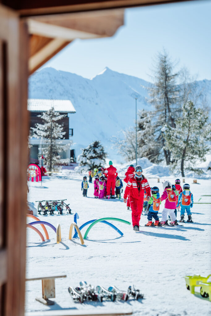 Photo en format portrait prise de jour en hiver. Nous pouvons voir le jardin d'enfants de l'ESF. Il y a deux groupes d'enfants entouré de 3 moniteurs de ski en train de donner un cours. 